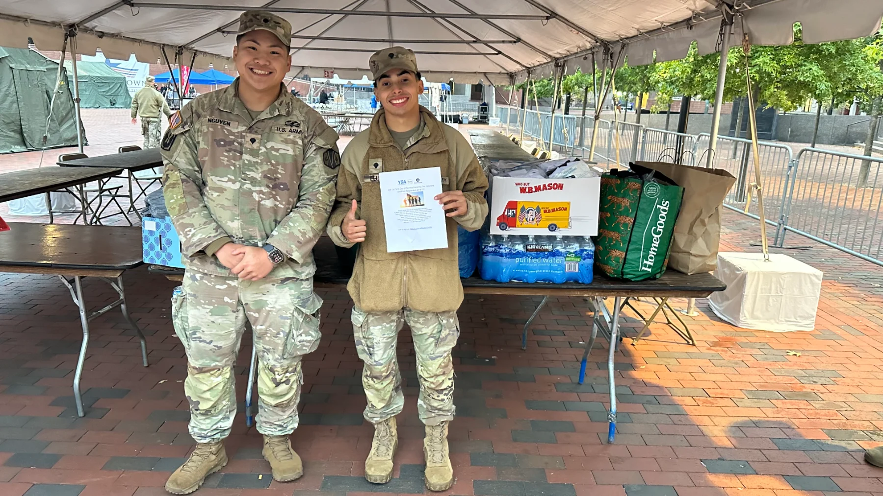 Two smiling U.S. Army service members standing fully in frame beside donated supplies